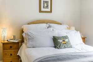 Ein ordentlich bezogenes Bett mit weißen Leinen und einem grünen Kissen mit einem Bienenmuster, ein Nachttisch aus Holz mit einer Lampe und einer Vase sowie ein gerahmtes Bild an der Wand.