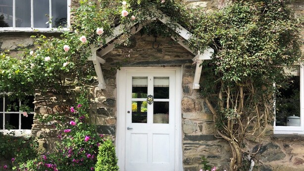 Ein Steinhaus mit einer weißen Tür, einer kleinen Veranda und einem Garten mit rosa Blüten.