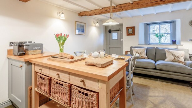 Une cuisine chaleureuse avec des poutres en bois, un coin repas avec un canapé et une fenêtre agrémentée d’une plante.