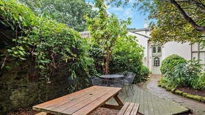 Ein Holztisch mit Bänken in einem Gartenbereich, eine Steinmauer, ein weißes Gebäude mit grüner Tür und ein kleiner runder Tisch mit Stühlen.