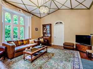 A spacious living room with a leather sofa, a coffee table, a television, and a large window with a view of greenery. - 6 Bed in Appleby-in-westmorland (oc-sz067) (Appleby-in-Westmoreland)