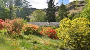 Ein Garten mit verschiedenen blühenden Pflanzen und Bäumen, einem gepflasterten Weg und einem Gebäude mit Schrägdach.