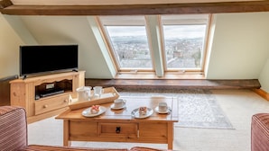 Ein gemütliches Zimmer mit Fernseher, Holzmöbeln, einem Esstisch mit Tellern und Tassen sowie Blick durch große Fenster.