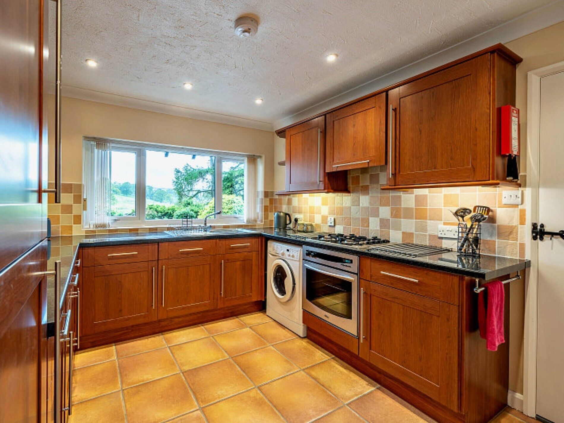 Une cuisine avec des meubles en bois, un lave-linge, une cuisinière et une fenêtre donnant sur un paysage verdoyant.