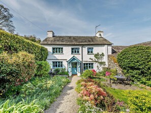Une maison à deux étages, avec un chemin de gravier menant à l’entrée, entourée d’une végétation luxuriante et d’un jardin.
