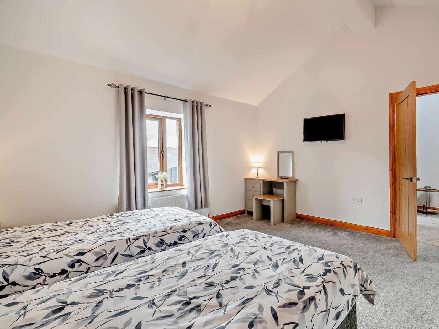 Ein Schlafzimmer mit zwei Betten, einem Fenster mit Vorhängen, einem Nachttisch mit Lampe, einem an der Wand befestigten Fernseher und einer Holztür.