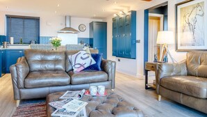 A modern living room with a leather sofa, ottoman, and a dining area with blue cabinets and a kitchen in the background. - 2 Bed in Stockport (oc-83789) (Mellor)