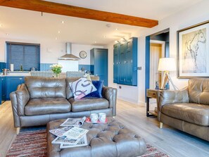 Ein modernes Wohnzimmer mit Ledersofa, Hocker und einem Essbereich mit blauen Schränken sowie einer Küche im Hintergrund.