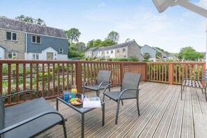 A wooden deck with outdoor furniture, a table with drinks and snacks, and a view of houses in the background. - 3 Bed in Falmouth (oc-42709) (Goldenbank)