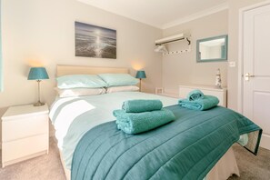 A neatly made bed with a teal comforter and pillows, a bedside lamp, a wall-mounted air conditioner, and a framed picture above the bed. - 3 Bed in Falmouth (oc-42709) (Goldenbank)