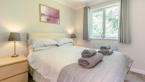 A neatly made bed with pillows, a bedside lamp, a nightstand, and a window with curtains. - 3 Bed in Falmouth (oc-42709) (Goldenbank)