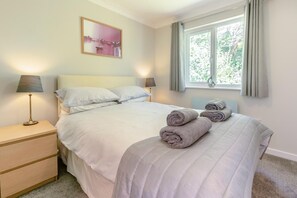 A neatly made bed with pillows, a bedside lamp, a nightstand, and a window with curtains. - 3 Bed in Falmouth (oc-42709) (Goldenbank)