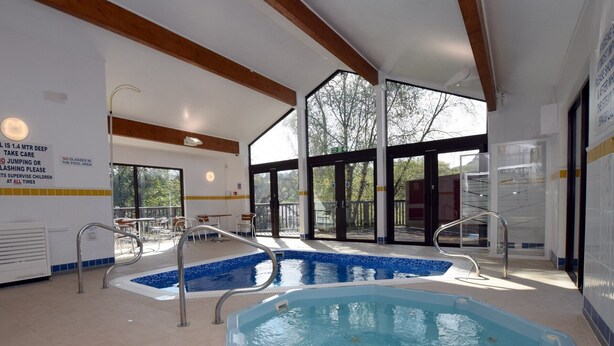 Una piscina interna con pareti in vetro trasparente, una vasca idromassaggio separata e un'area a bordo piscina con tavoli e sedie.