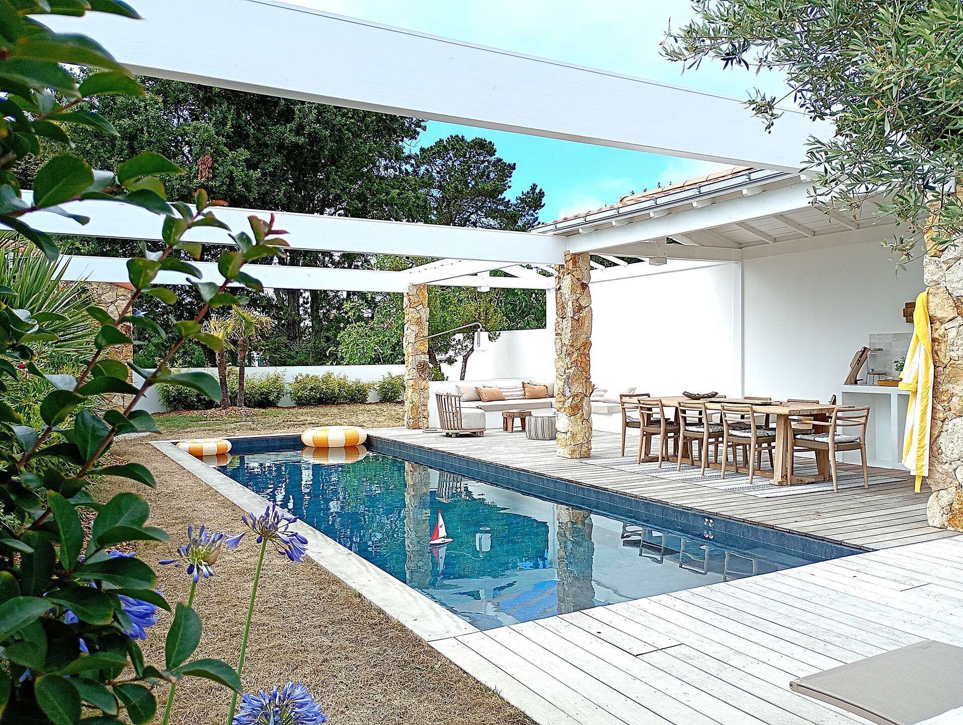 Un área de piscina con terraza de madera, zona de estar con sillas y una mesa, y un pilar de piedra.