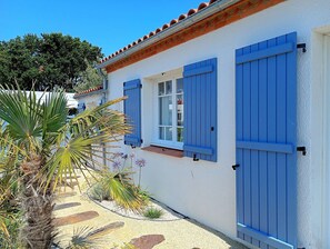 Ein weißes Gebäude mit blauen Fensterläden, einem Ziegeldach und einem Garten mit Palmen und blühenden Pflanzen.