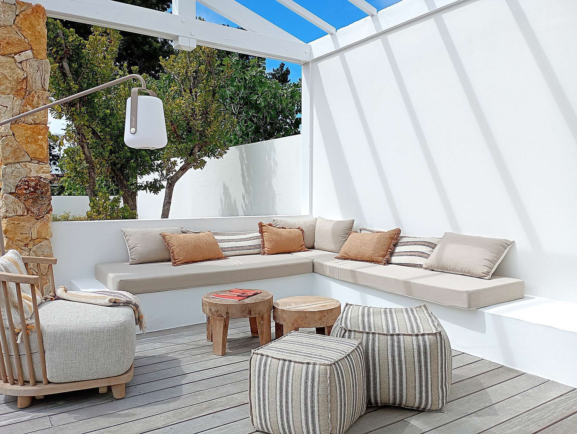 A covered outdoor seating area with a white wall, stone pillar, and a view of trees.