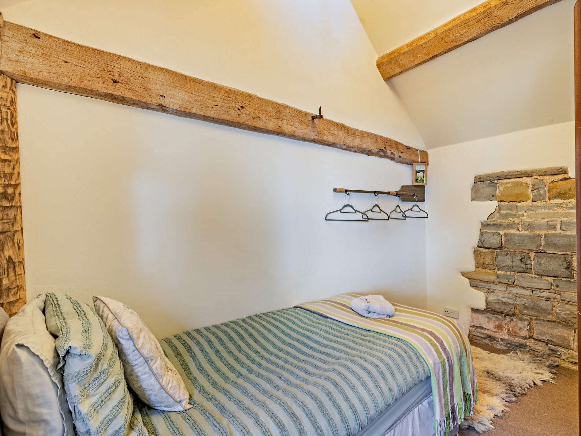 Een slaapkamer met een bed, houten balken, een stenen muur en een kapstok met ophanghaakjes.