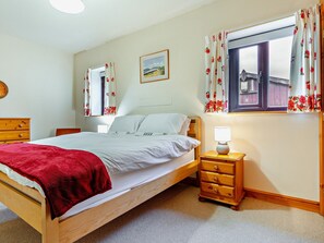 Ein Schlafzimmer mit einem hölzernen Bett, einem Nachttisch mit Lampe, einer Kommode, einem Fenster mit floralem Vorhang und einem gerahmten Bild an der Wand.