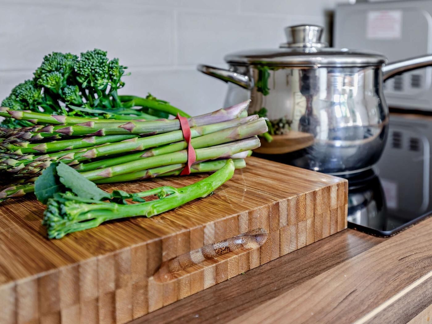 Una tabla de madera con espárragos y brócoli, una olla de acero inoxidable y una encimera de cocina.