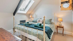Ein Schlafzimmer mit schrägen Wänden, einem Bett mit Metallgestell, zwei Nachttischen mit Lampen, einem Heizkörper und einem kleinen Holztisch.
