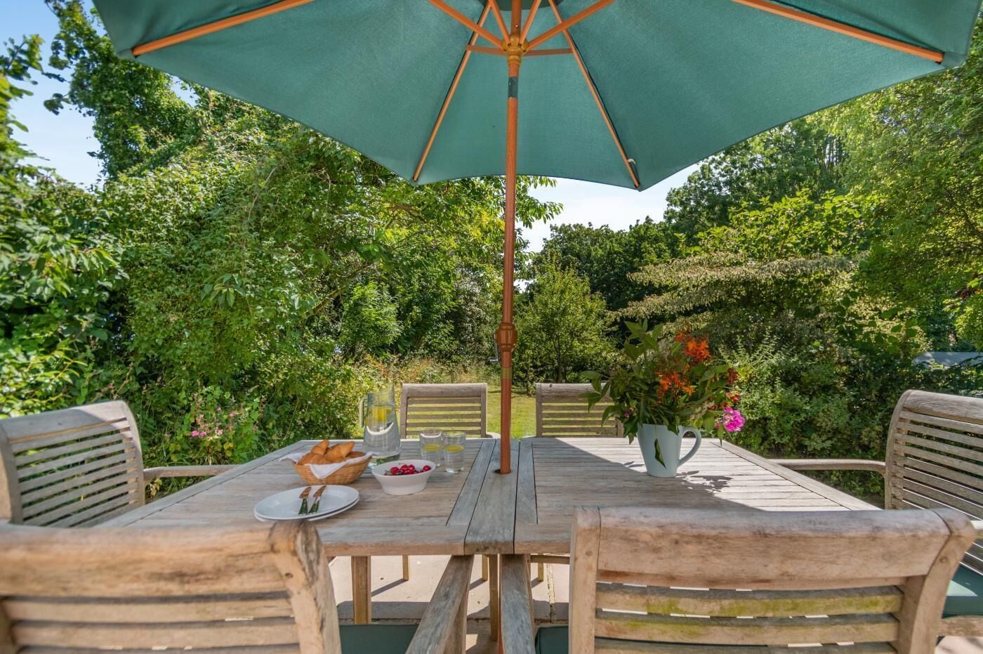 An outdoor wooden table with chairs, a green umbrella, and a pitcher of juice.