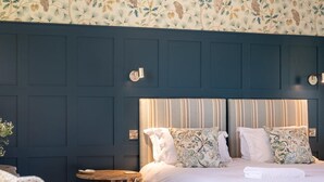 Une chambre avec un mur lambrissé bleu foncé, deux lits dotés de têtes de lit à rayures, des coussins à motifs et une petite table de chevet en bois.