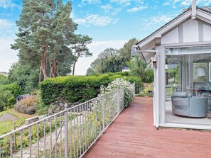 Ein überdachter Außenbereich mit Holzterrasse, Metallgeländer und Blick auf einen Garten mit Bäumen und Sträuchern.