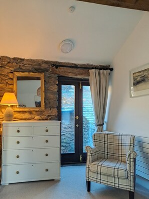 A room with a stone wall, a white dresser with drawers, a plaid sofa, a mirror, and a door leading outside. - 1 Bed in Patterdale (oc-sz154) (Glenridding)