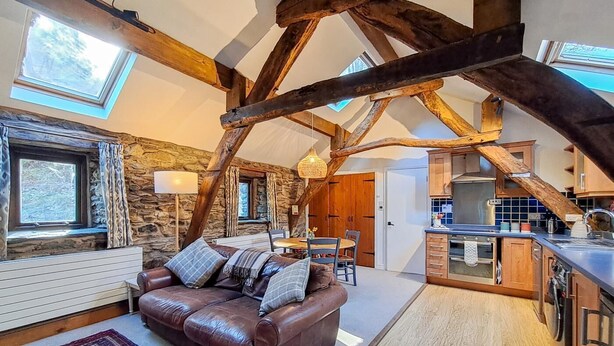 Un soggiorno in stile loft con travi a vista in legno, una parete in pietra, un divano in pelle, un tavolo da pranzo e una cucina con mobili in legno e un'ampia finestra.