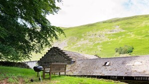 Un edificio in pietra con tetto spiovente, una panchina di legno e un barbecue in una zona erbosa con una collina sullo sfondo.
