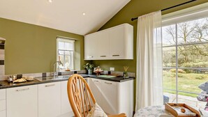 Une cuisine avec des meubles blancs, une chaise en bois, une table à manger recouverte d’une nappe à motifs floraux et une fenêtre donnant sur l’extérieur.