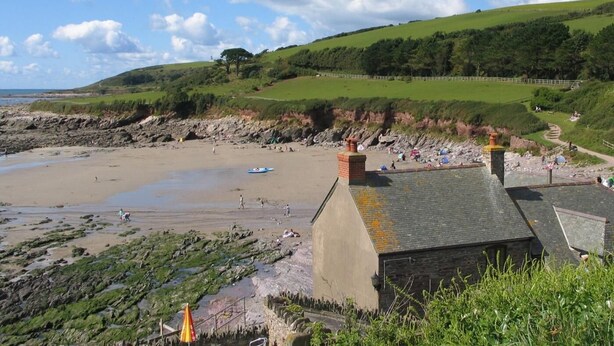 Una playa costera con costas rocosas, un pequeño edificio de techo inclinado y un camino que lleva a la playa.