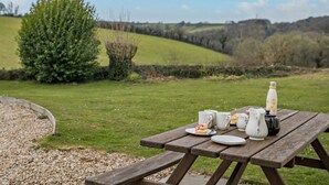 Una mesa de picnic con una botella, vasos y un plato de comida sobre un campo cubierto de césped, con vistas a colinas ondulantes.