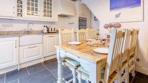 A kitchen with white cabinets, a wooden dining table, and a blue painting on the wall. - 3 Bed in Bideford (oc-57804) (Appledore)