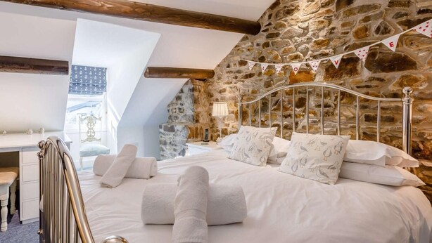 A bedroom with a stone wall, a sloped ceiling, a bed with white bedding, and a desk with a chair. - 3 Bed in Bideford (oc-57804) (Appledore)