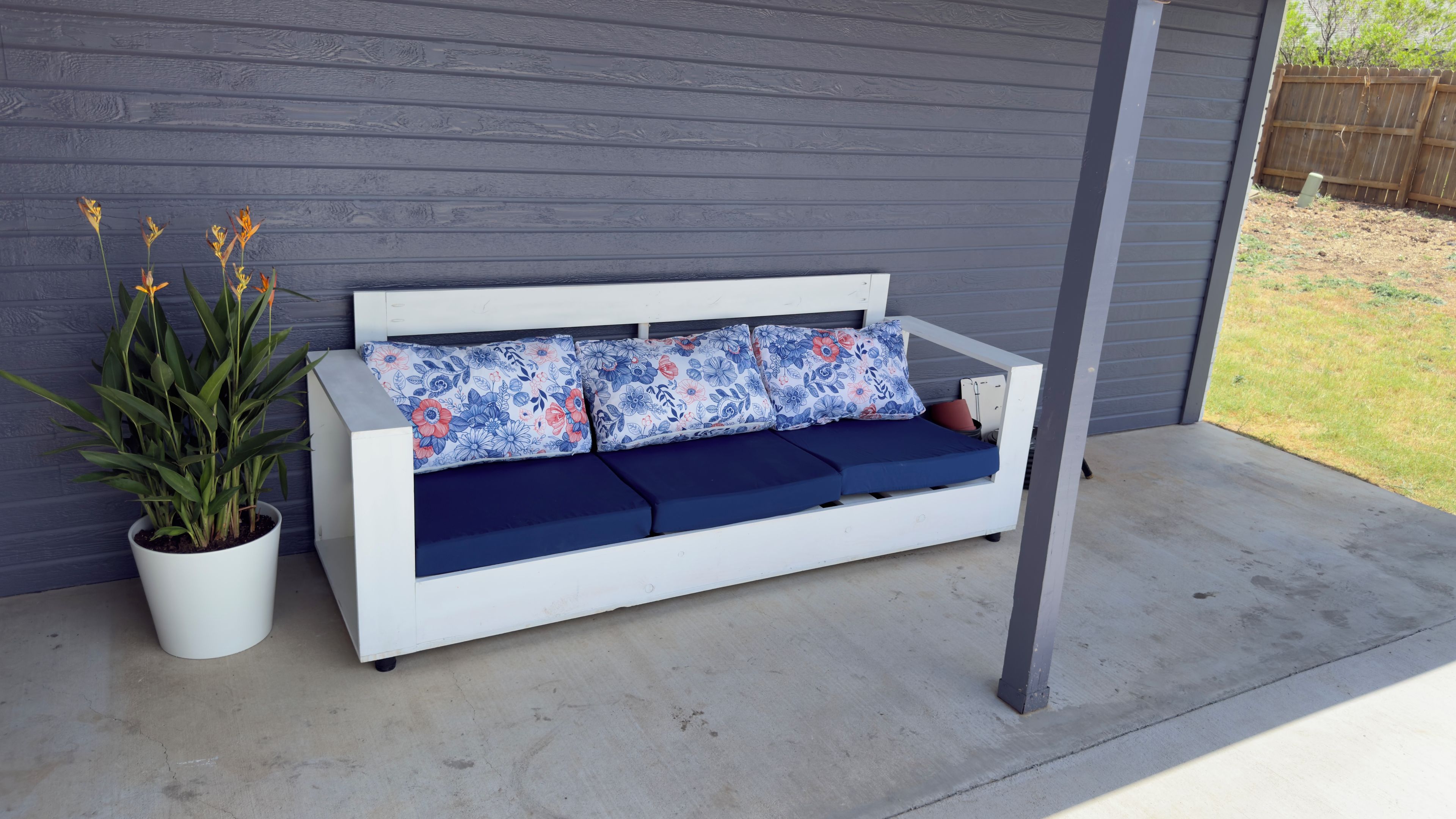 A white outdoor sofa with blue cushions and floral patterns, positioned against a grey wall with a potted plant beside it.