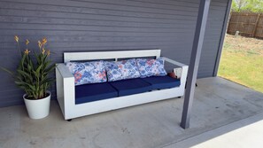 Ein weißes Outdoor-Sofa mit blauen Kissen und floralen Mustern, platziert vor einer grauen Wand mit einem Blumentopf daneben.