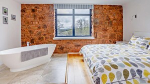 Een moderne badkamer met een vrijstaand bad, een groot raam met uitzicht op bomen en een bed met een gestreept dekbed.