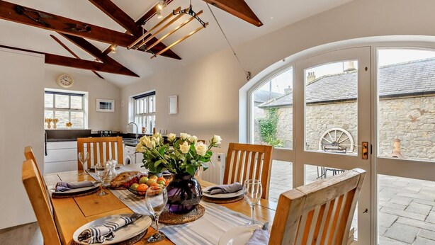 A dining area with a wooden table set for four, a vase with flowers, and a view of a stone building through a glass door. - 3 Bed in Alnwick (oc-94195) (Christon Bank)