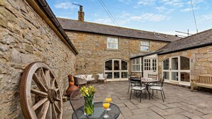 A stone building with a courtyard, outdoor seating, and a large wooden wheel. - 3 Bed in Alnwick (oc-94195) (Christon Bank)