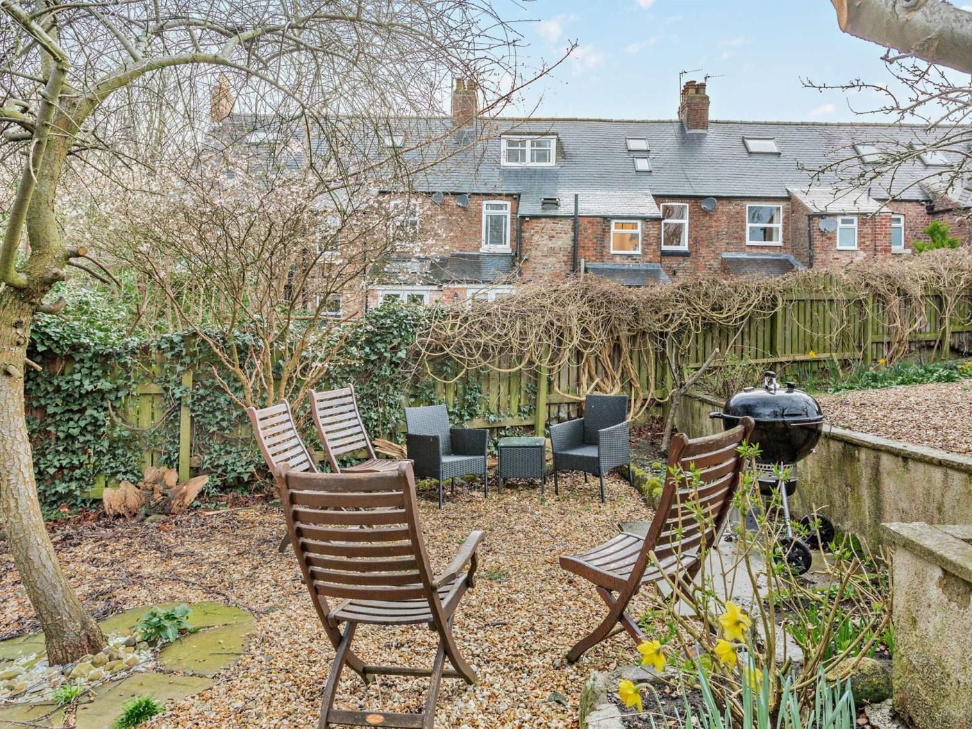 Un patio trasero con una parrilla, dos sillas de madera, una mesa y un jardín con plantas y un árbol.