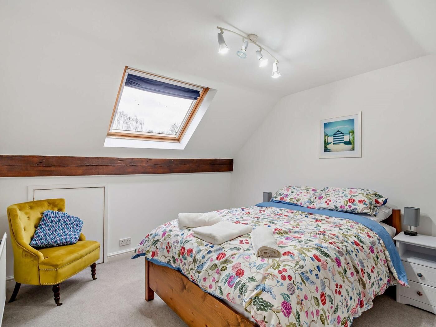 Een slaapkamer met dakraam, een bed met bloemenbedrukte lakens, een gele fauteuil en een nachtkastje met een lamp.