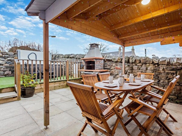 An outdoor patio with a stone wall, a wooden table with chairs, and a barbecue grill. - 3 Bed in Grantown-on-spey (oc-94514) (Grantown-on-Spey)