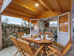 An outdoor wooden dining area with a stone wall backdrop, a round table set for four, and a cozy interior visible through an open door. - 3 Bed in Grantown-on-spey (oc-94514) (Grantown-on-Spey)