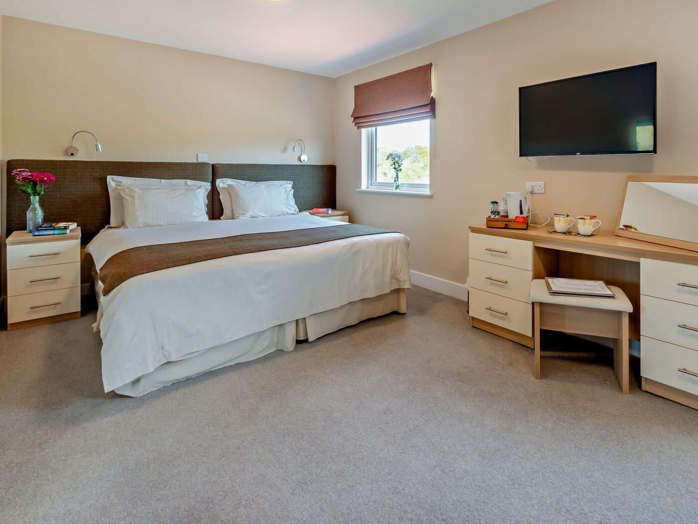 Une chambre à coucher comprenant un lit, des tables de chevet, un bureau, une chaise, un téléviseur et une fenêtre donnant sur un paysage agréable.