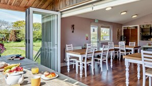 A dining area with wooden tables and chairs, a view of the outdoors, and a kitchen in the background. - 5 Bed in Ventnor (oc-94494) (Godshill)