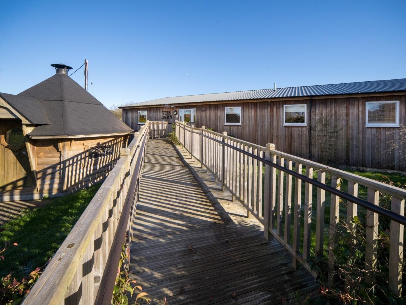 Een houten looppad dat leidt naar een landelijk gebouw genaamd 'The Barn', met een metalen dak en houten bekleding.