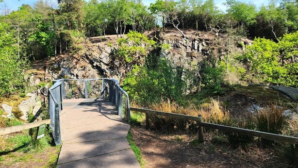 Un chemin en béton mène à un pont métallique qui enjambe un terrain rocheux recouvert d’une végétation luxuriante.