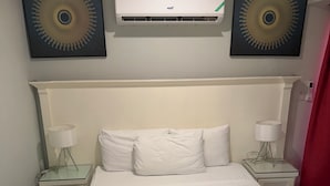 A hotel room with a bed, white bedding, two bedside lamps, an air conditioning unit, and two wall-mounted decorative items. - Relax in luxury and sleep in peace. (Discovery Bay)