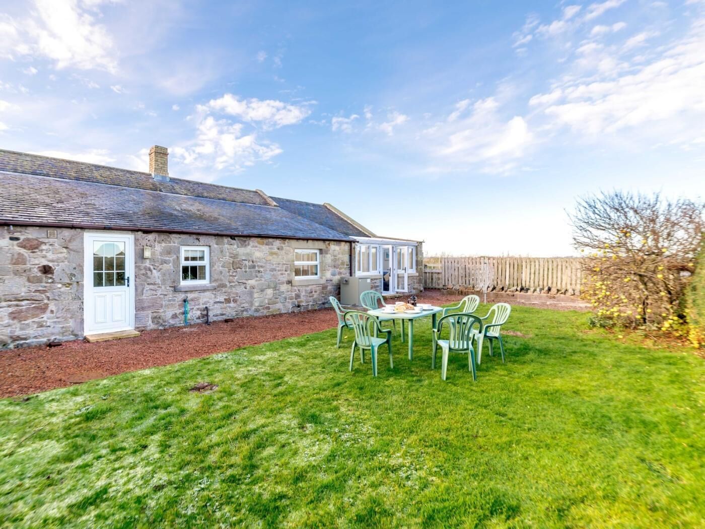 A stone house with a white door, a small porch, and a grassy yard with a table and chairs.